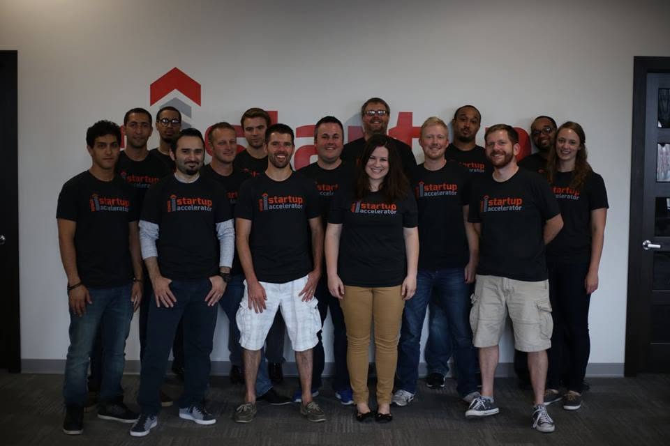 A group of participants wearing Iowa Startup Accelerator shirts, gathered in front of the accelerator wall